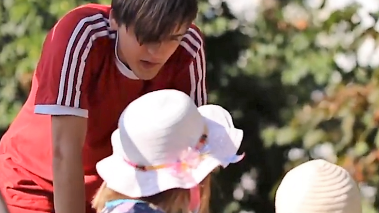 Junger Mann in rotem T-Shirt schiebt zwei kleine Mädchen mit Sonnenhüten in einer Nestschaukel an 