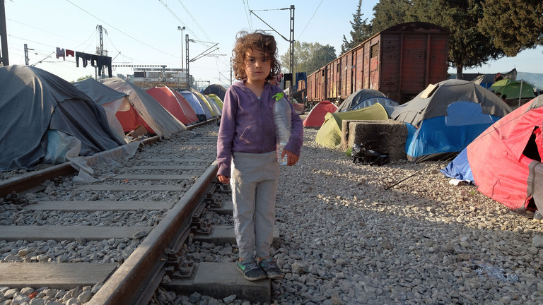Ca. 4jähriges Mädchen an einem Bahngleis, das durch ein griechisches Flüchtlingslager fürht, rechts und links stehen eingestaubte Igluzelte 