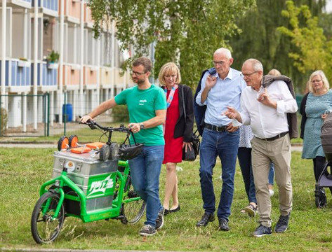 Ein Mann schiebt ein Lastenfahrrad über eine Grünfläche, dahinter eine Frau und zwei Männer um die 60 Jahre im Gespräch, im Hintergrund sanierte DDR-Plattenbauten
