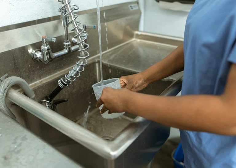 Mensch in blauer Krankenhauskleidung spült einen Becher in Edelstahl-Becken