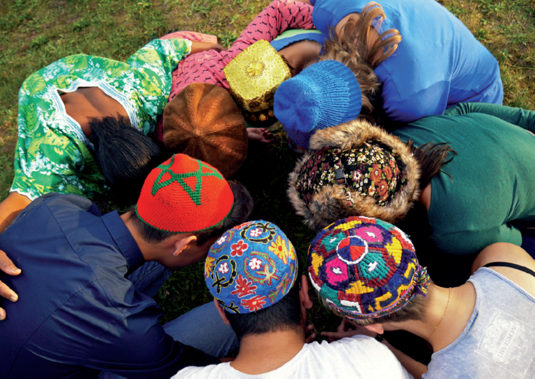 Junge Menschen mit verschiedenen bunten Kopfbedeckungen, darunter eine mit dem Davidstern, stecken die Kopfe zusammen